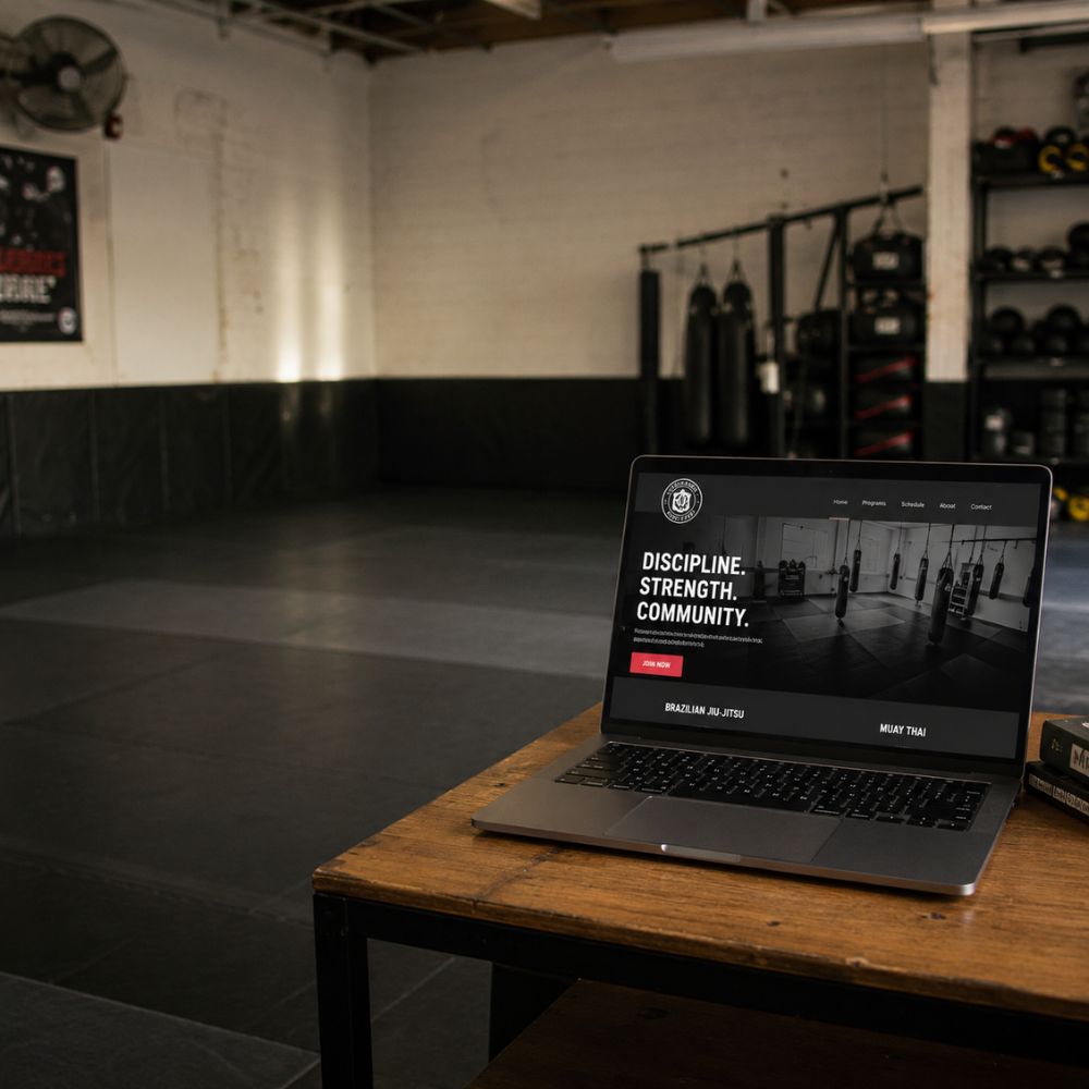 Laptop displaying a martial arts gym website with the headline 'Discipline. Strength. Community.' on a wooden table inside a boxing gym